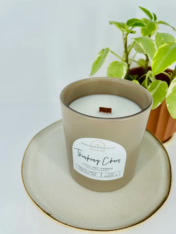 A brown coconut soy candle labeled "Tahitian Citrus" with a wooden wick, discovered as one of January 2025's coveted FINDS, rests on a ceramic plate beside a potted plant, evoking the vibrant spirit of Memphis.